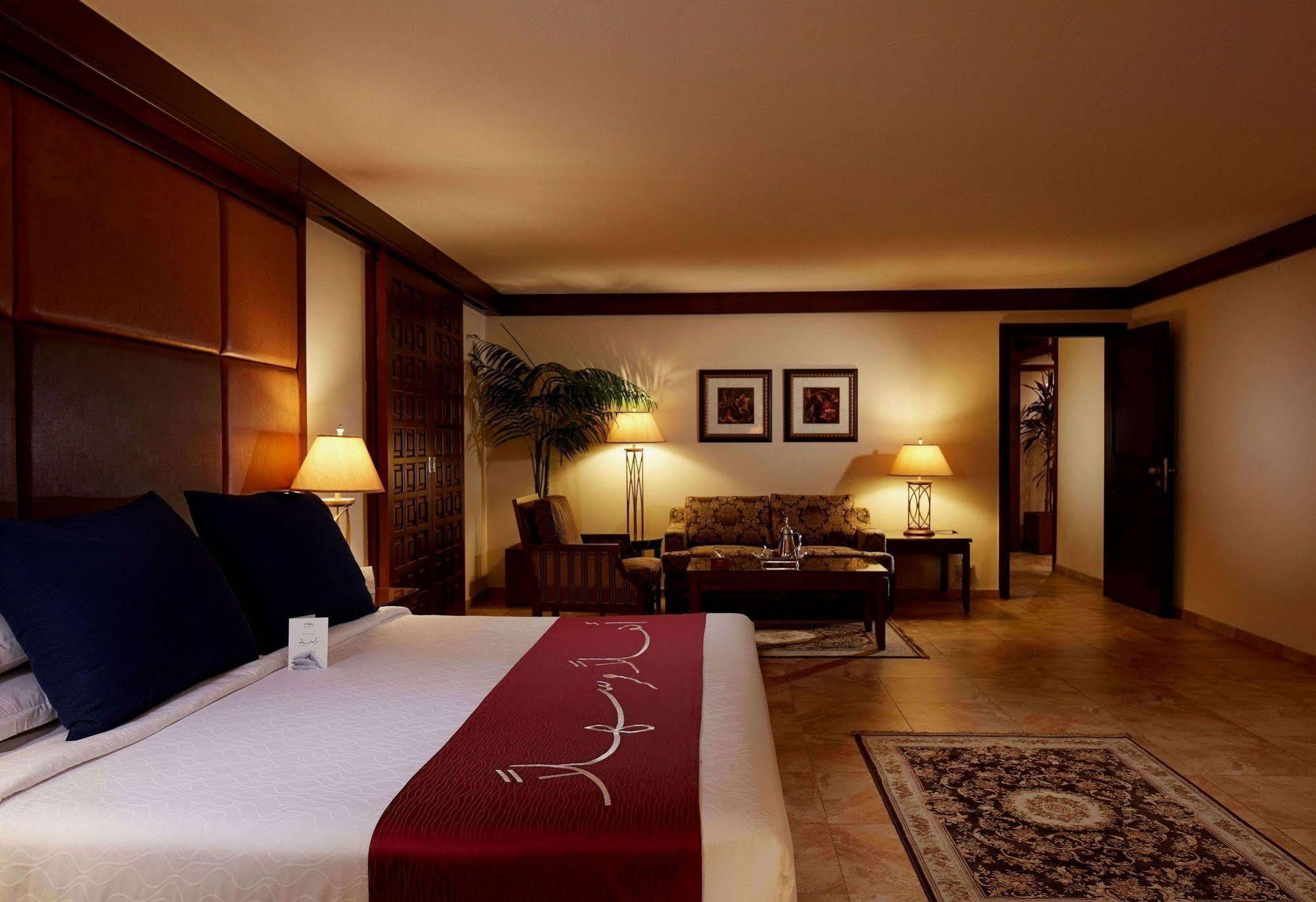 Makarem Ajyad Makkah Hotel La Meca Exterior foto The photo depicts a cozy hotel room with a warm and inviting atmosphere. It features a large bed with dark blue pillows and a maroon throw blanket. On either side of the bed, there are lamps providing soft lighting. In the background, a small seating