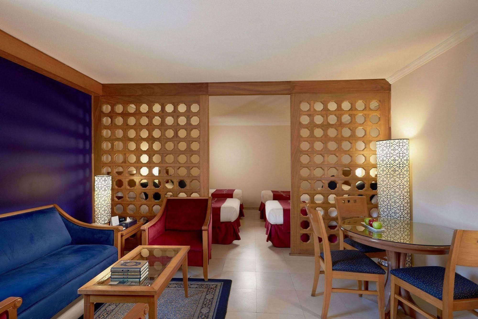 Makarem Ajyad Makkah Hotel La Meca Exterior foto The photo shows a well-furnished hotel room or suite. On the left side, there is a seating area featuring a blue sofa and a small coffee table, as well as a couple of decorative items. The room is separated by a decorative wooden partition with circu