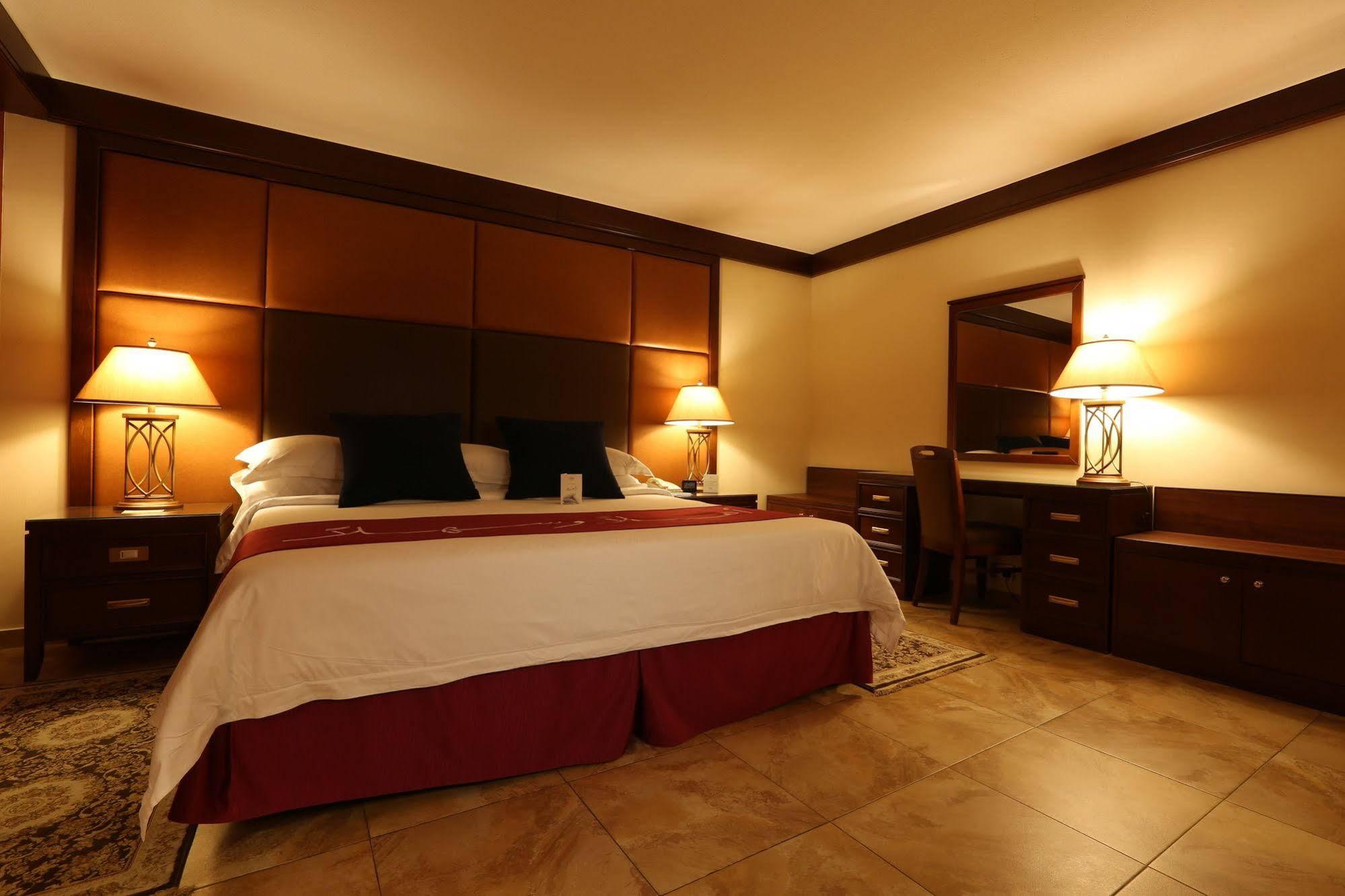 Makarem Ajyad Makkah Hotel La Meca Exterior foto The photo shows a well-appointed hotel room with a cozy and inviting atmosphere. It features a large bed with a white comforter and dark pillows, positioned against a wooden headboard. There are two bedside lamps providing soft lighting on either sid