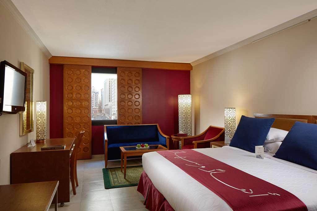 Makarem Ajyad Makkah Hotel La Meca Habitación foto The photo showcases a hotel room featuring a modern design. In the center, there's a large bed with white linens and a decorative red runner. On either side of the bed, there are wooden nightstands with lamps. A blue sitting area is visible, composed