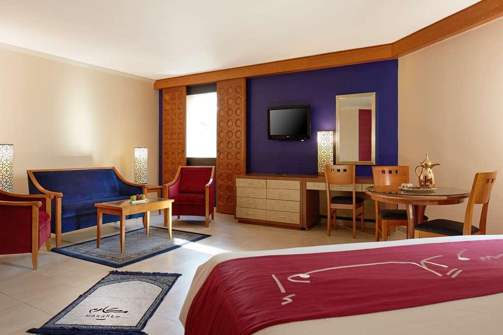 Makarem Ajyad Makkah Hotel La Meca Habitación foto The image shows a hotel room with a modern, comfortable design. The room features a large bed with a red and white bedspread, a seating area that includes a blue sofa, two red chairs, and a wooden coffee table. There's a flat-screen TV mounted on the
