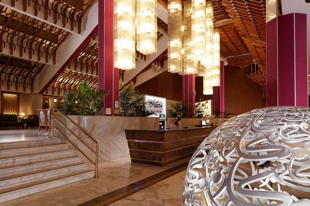 Makarem Ajyad Makkah Hotel La Meca Interior foto The photo depicts a luxurious hotel lobby. It features a grand entrance with a staircase leading up to a reception area. The lobby has high ceilings adorned with striking, modern light fixtures that hang down, providing a warm ambiance. The walls are
