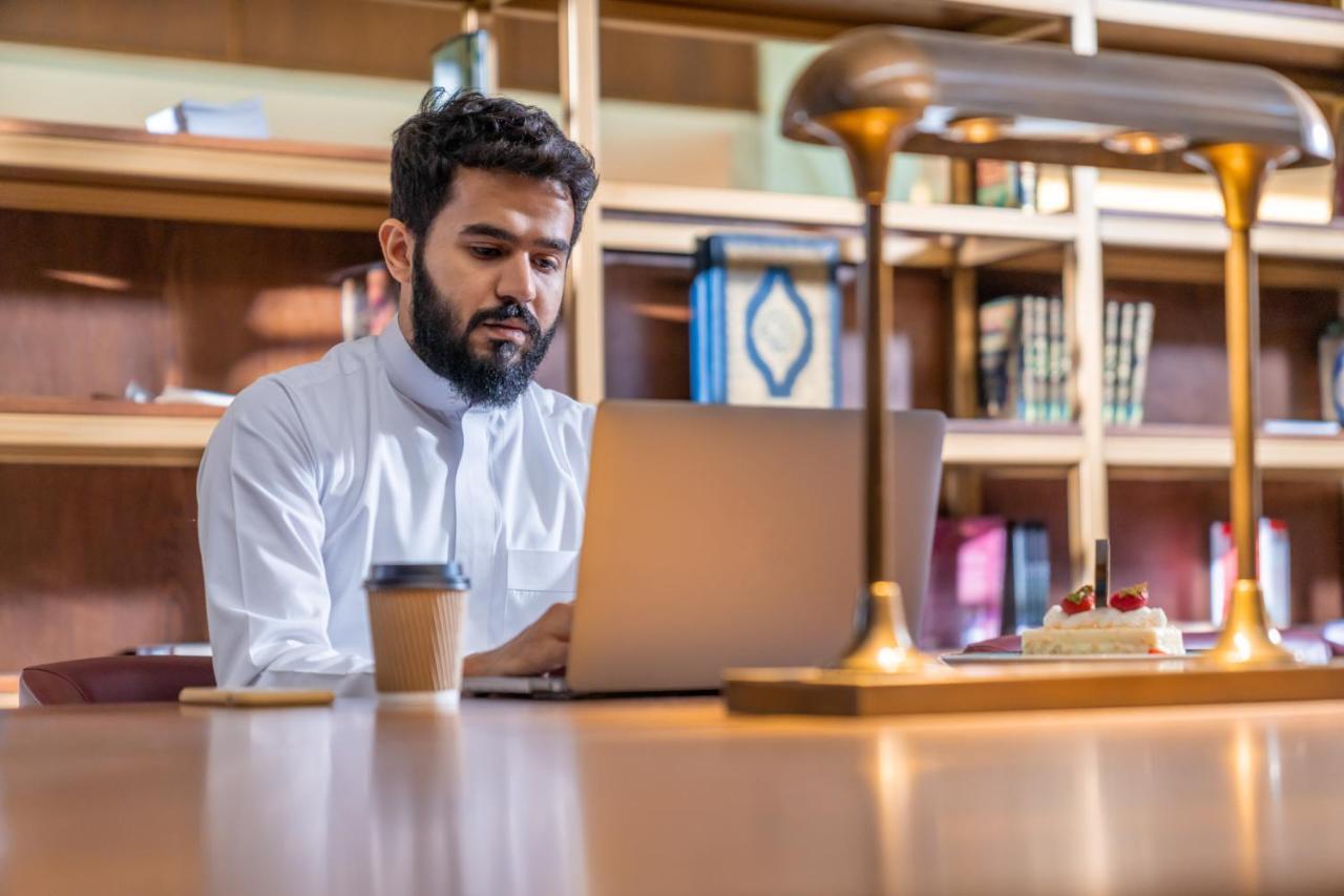 Makarem Ajyad Makkah Hotel La Meca Exterior foto The photo shows a man sitting at a desk in a library or a study area. He is wearing a white traditional garment and is focused on working on a laptop. In front of him, there is a cup of coffee and a small cake decorated with strawberries. The backgro