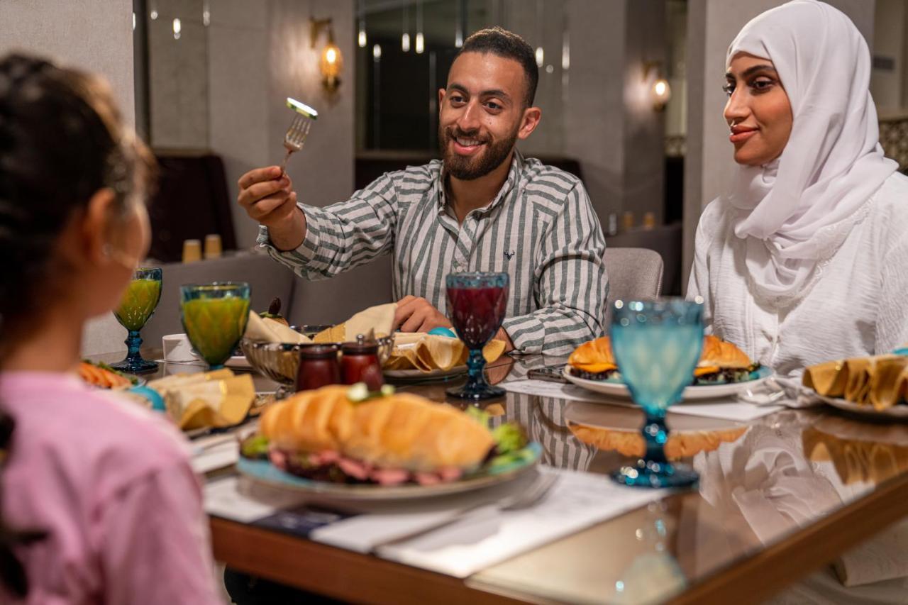 Makarem Ajyad Makkah Hotel La Meca Exterior foto The photo shows a dining scene with three people enjoying a meal together. A man is seated at the table, smiling and holding a utensil, possibly ready to serve or share food. He is dressed in a striped shirt. Across from him is a woman wearing a whit