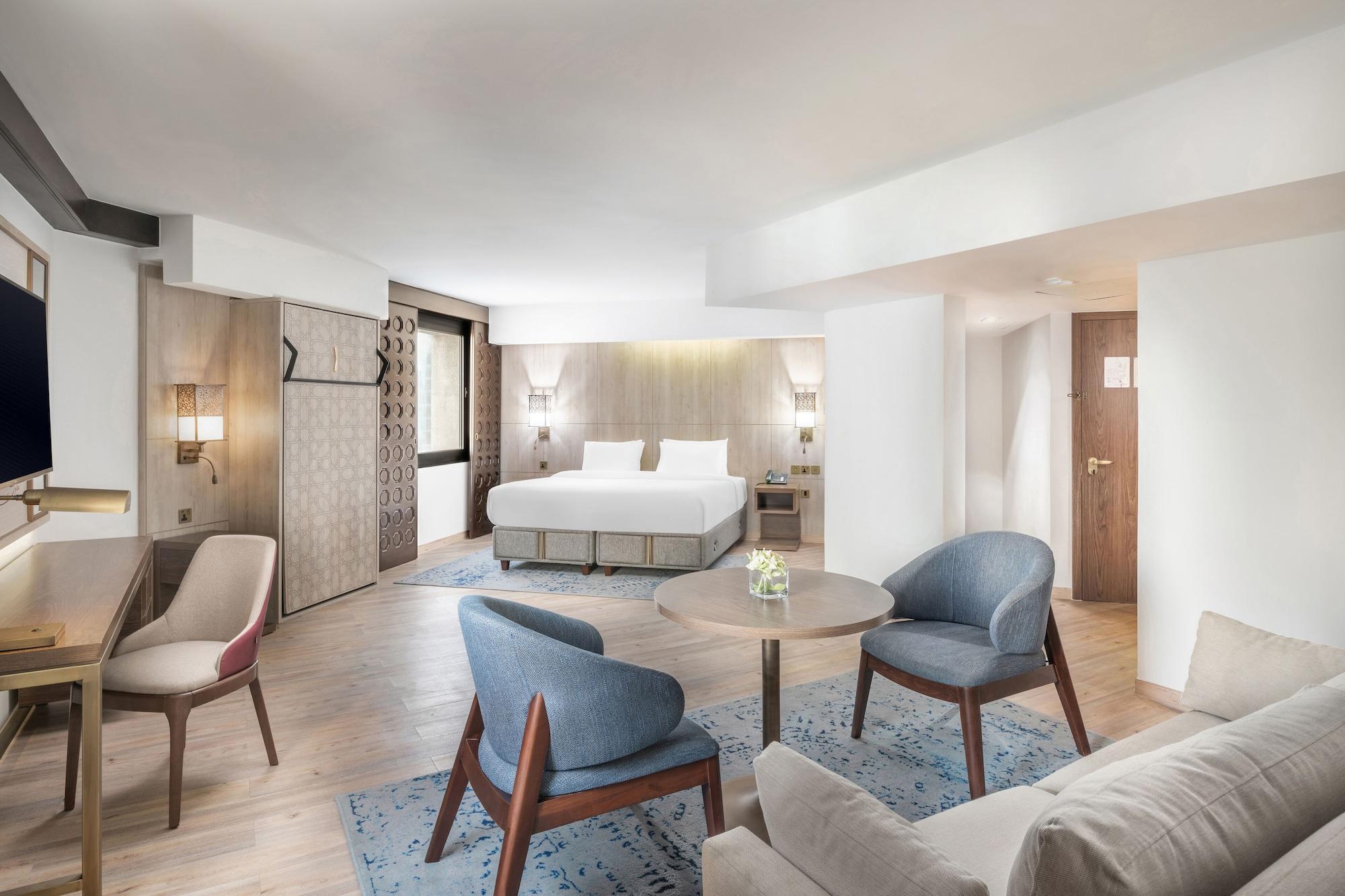 Makarem Ajyad Makkah Hotel La Meca Exterior foto The photo shows a modern hotel room with a spacious layout. It features a large bed with a simple headboard against a light-colored wall. There are two soft chairs in light blue and beige positioned around a small round table. The flooring is wooden,