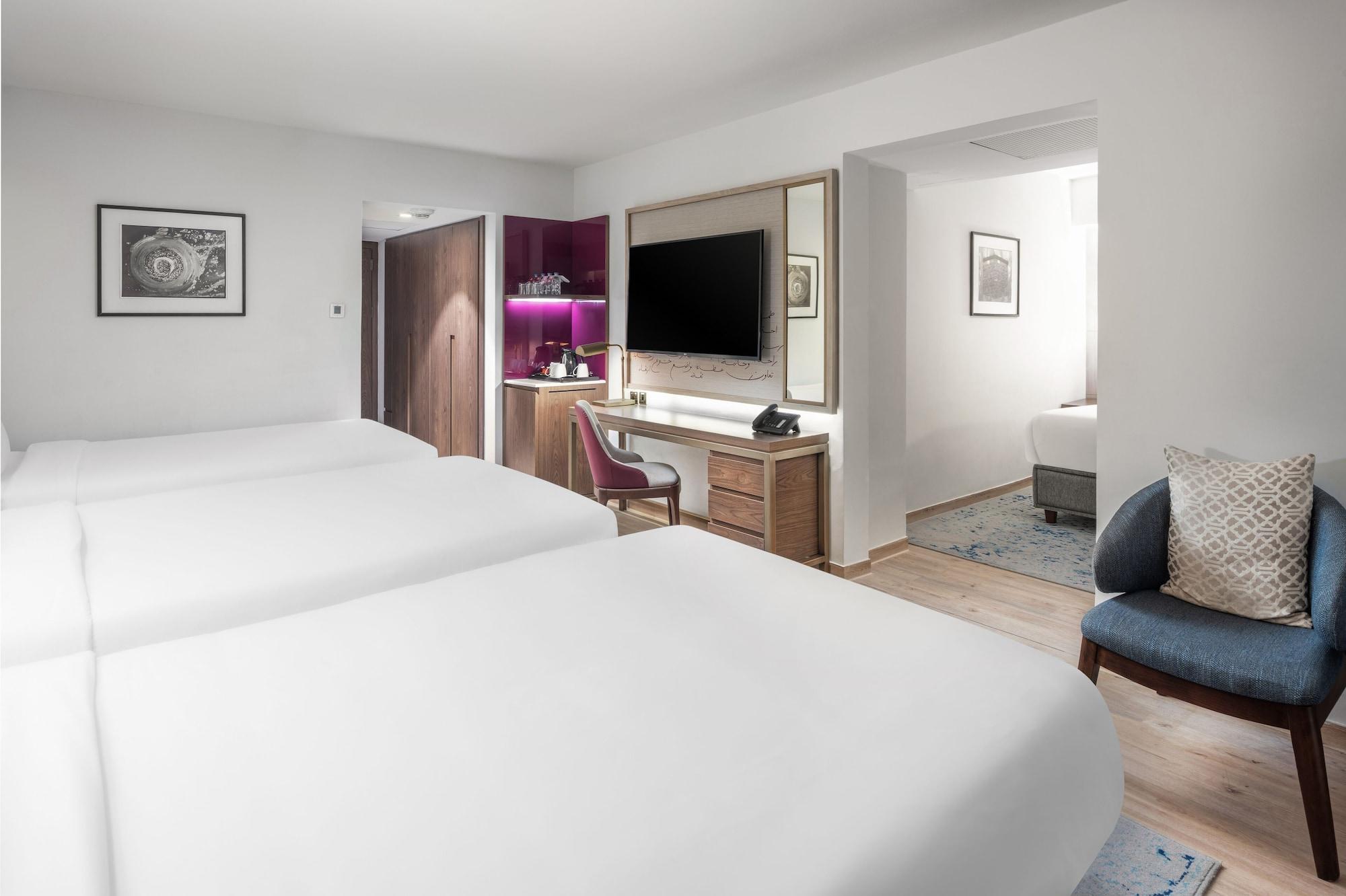 Makarem Ajyad Makkah Hotel La Meca Exterior foto The photo shows a modern hotel room featuring two white beds with smooth linens. The room has a minimalistic design with light-colored walls and wooden flooring. On one side, there is a desk and chair next to a television mounted on the wall, along w