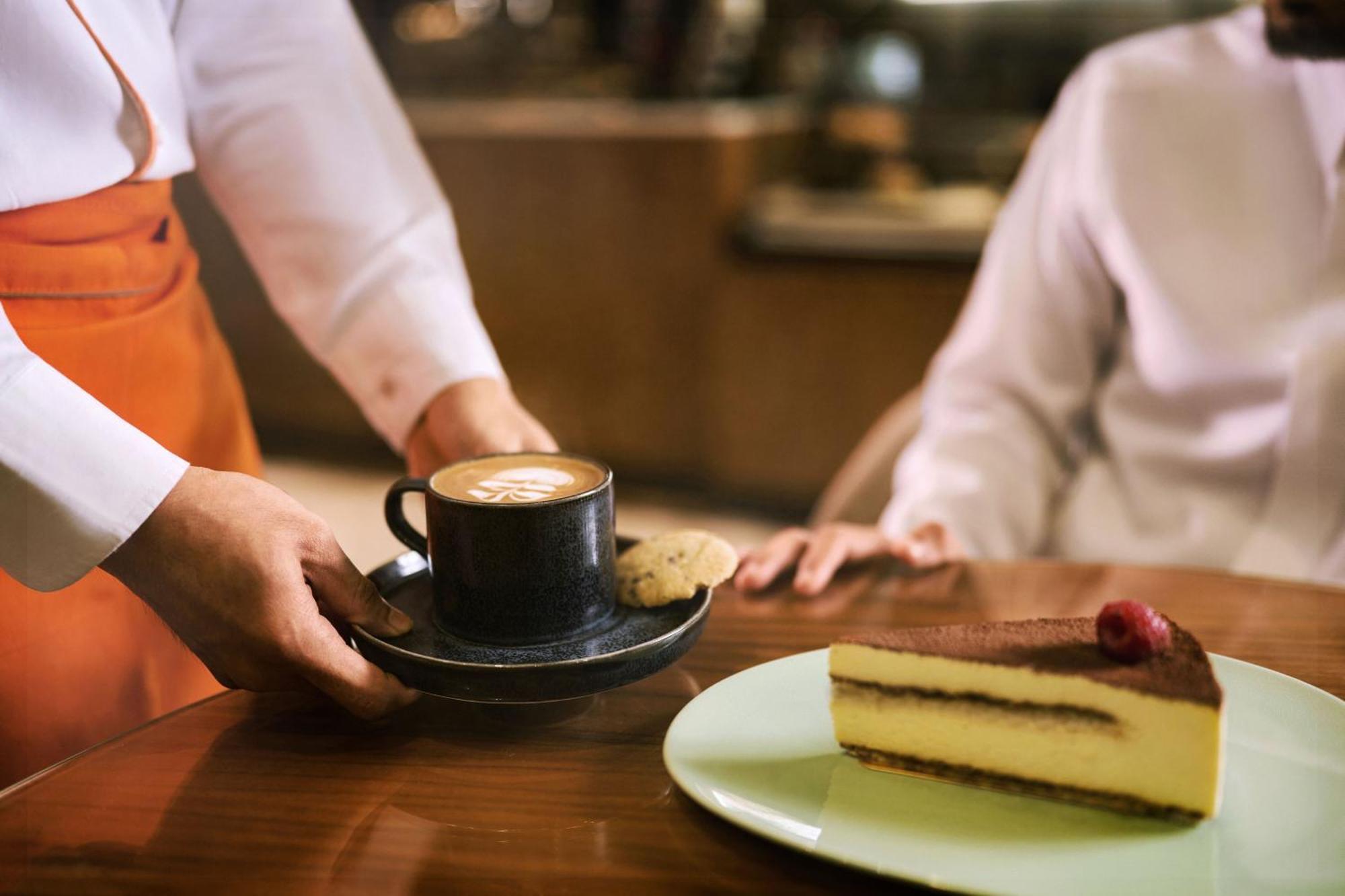 Makarem Ajyad Makkah Hotel La Meca Exterior foto The photo shows a hand presenting a cup of coffee on a saucer, accompanied by a cookie. In the foreground, there is a slice of cake on a light green plate, garnished with a raspberry. The setting appears to be a café or restaurant, with a blurred bac