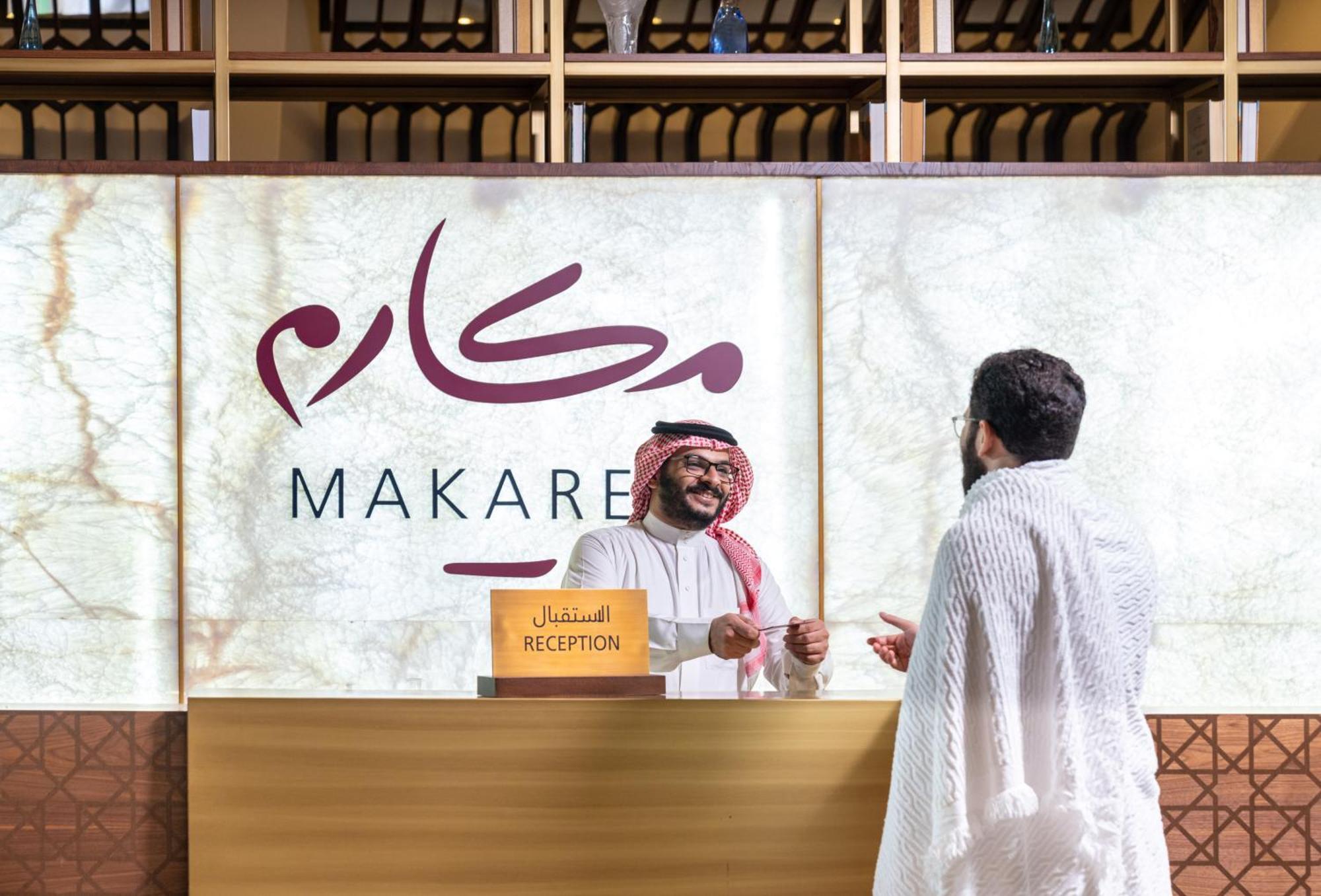 Makarem Ajyad Makkah Hotel La Meca Exterior foto The photo depicts a reception area in a hotel or establishment named "Makarem." In the image, a receptionist, dressed in traditional attire, is smiling and interacting with a guest who is wearing a white robe, commonly known as a thobe or ihram. The 