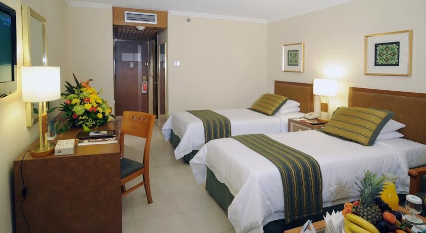 Makarem Ajyad Makkah Hotel La Meca Exterior foto The photo shows an interior of a hotel room featuring two twin beds with white linens and green decorative accents. The walls are painted in a light beige color, and there is a small table next to one of the beds. A desk with a chair is positioned ne